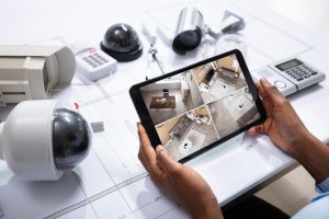 An Elevated View Of Person Watching Footage On Digital Tablet With Security Equipment On Blueprint